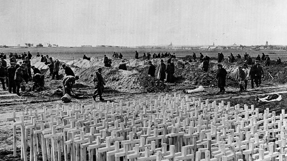 Schwarz-weiß-Foto, im Vordergrund Dutzende weiße Kreuze im Boden, dahinter Menschen mit Schaufeln, die weitere Gräber ausheben