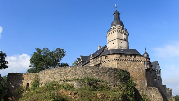 Zu sehen ist die Burg Falkenstein, leicht von unten fotografiert. Vorne sieht man Teile der Burgmauer. Der Himmel ist blau.