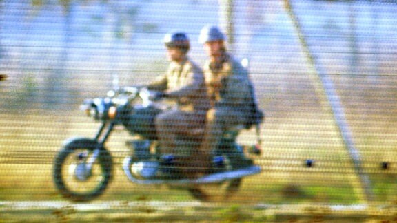 DDR Grenzer hinterm Grenzzaun auf einem Motorrad mit Maschinenpistole