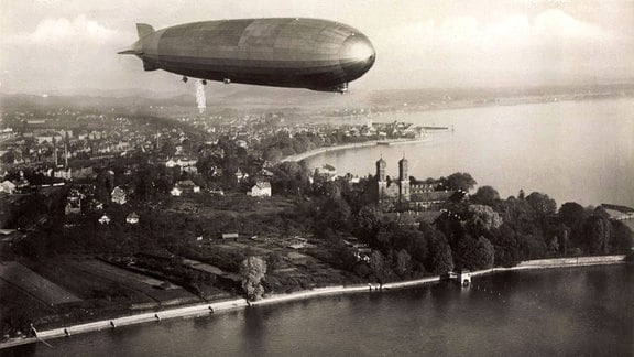 Ein Zeppelin fliegt über eine Stadt an einem See.