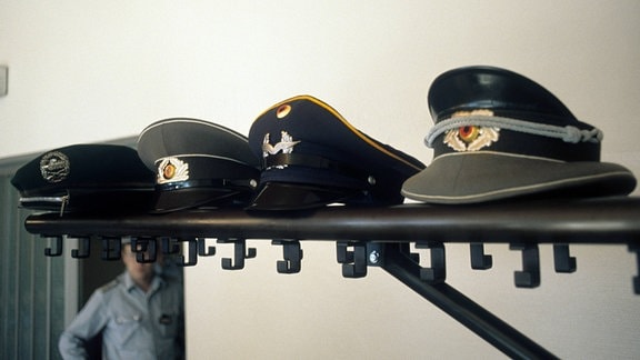 NVA- und Bundeswehr-Mützen liegen auf einem Regal.