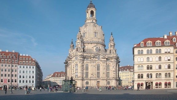 Die Sandsteinquader der wiederaufgebauten Frauenkirche zu Dresden strahlen im Sonnenschein. Viele Menschen flanieren im sanierten Altstadtzentrum rund um die Frauenkirche.