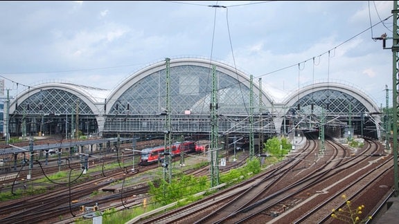 Hauptbahnhof zu Dresden. Im Vordergrund Gleise. Verlaufen nach hinten. Darüber drei Glasbögen. Mittlerer größer als die anderen beiden links und rechts davon. Ein Zug verlässt den Hauptbahnhof; einer fährt gerade ein.