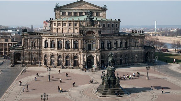 Semperoper in Dresden, vor dem Gebäude ein Platz mit Menschen und dem Reiterstandbild, das König Johann von Sachsen darstellt.