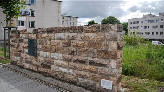 Am Grundstück der ehemaligen Dresdner Stasi-Zentrale in der Bautzner Straße steht ein Stück Mauer mit Gedenktafel. Dahinter Wiese und Häuser.
