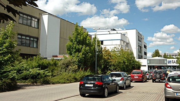 Standort des Halbleiterproduzenten X-Fab in Erfurt in der Haarbergstraße. Mehrteiliges Firmengebäude mit Flachdächern, beiger und weißen Fassaden. Im Hintergrund ist der Eingang mit dem Schriftzug XFab zu sehen. Vor den Gebäuden gitbes einen schmalen Grünstreifen mit Büschen und Bäumen. Mehrere Autos parken.