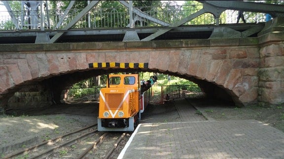 Kleine Parkeisenbahn mit orangefarbener Lok fährt unter einer Brücke hindurch.