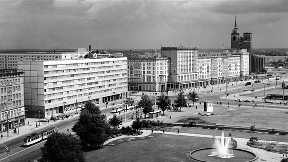 Die Wilhelm-Pieck-Allee in Magdeburg im jahr 1968, Aufnahme in Schwarz-weiß. Auf der linken Seite steht der sogenannte Blaue Bock, ein Arbeiterwohnheim mit blaugekachelter Fassade. Hinten rechts ist die Johanniskirche zu sehen. Deren rechter Turm hat keine Spitze. Die rechte Seite der Allee ist unbebaut, vorne befindet sich ein Springbrunnen in Betrieb.
