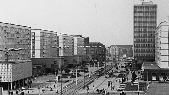 Die Karl-Marx-Straße in Magdeburg in der DDR-Zeit. Aufnahme in Schwarz-weiß. Plattenbauten säumen die Fußgängerzone mit Straßenbahnverkehr. Hinten rechts steht das Hochhaus "Haus des Lehrers". Davor ist das Gebäude mit dem Restaurant "Ratswaage" zu sehen, im Vordergrund die Marietta-Milchbar mit Freisitz. Dei Straße ist großflächig mit Gehwegplatten belegt. Heute heißt die Straße Breiter Weg.