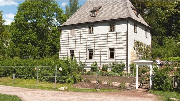 Zweistöckiges Haus mit Holzleisten auf der grau gestrichenen Fassade im Grünen, Bauzaun vor dem Haus.