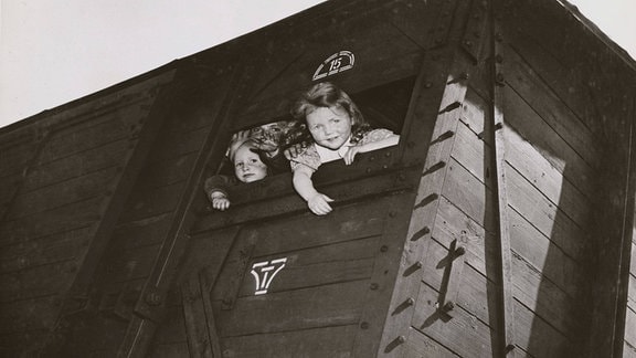 Historische schwarz-weiß-Aufnahme. Aus einem Zugwaggon ohne Fenster gucken drei Kinder, etwa vier Jahre alt, aus einer heruntergelassenen Klappe heraus