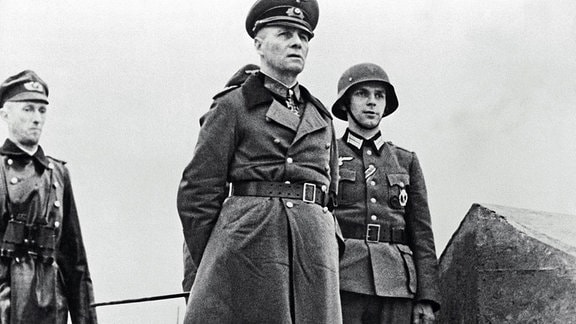 General Rommel inspiziert den Küstenschutz mit einer Gruppe deutscher Offiziere. Vor ihm ist ein MG-Schütze.