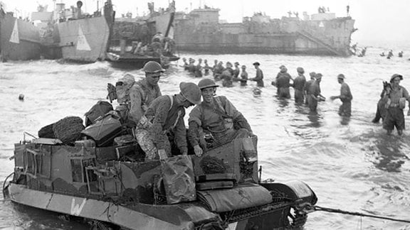 Ein Universal-Transporter wird an Land geschleppt, während im Hintergrund Truppen Munition aus einem Landungsboot entladen, 10. Juli 1943.
