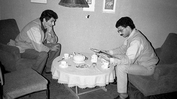Zwei Männer, einer skeptisch die Hand am Kinn, der andere in einem Buch lesend, sitzen in Sesseln an einem runden flachem Tisch auf welchem Teetassen, zwei Kannen und Gebäck stehen.