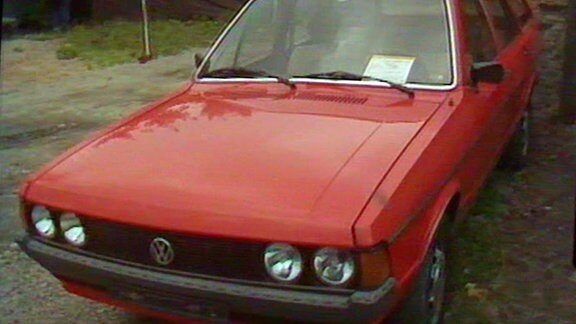 Vor einem Privatgrundstück steht ein alter, roter VW Passat mit einem Verkaufsschild hinter der Frontscheibe.