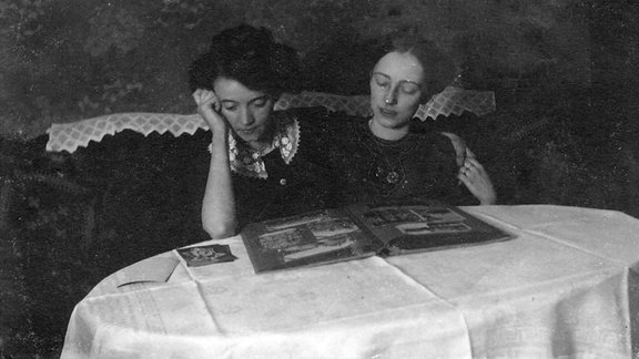 Schwarz-Weiß-Foto: Zwei junge Frauen sitzen an einem Tisch und schauen in ein Album.