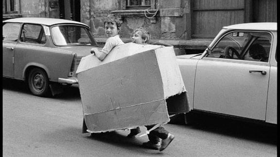 zwei kleine Jungs laufen auf der Straße mit einem Pappkarton herum und Spielen Auto.Dahinter stehen auch zwei Pappkartons, jedoch richtige Autos. Trabbis.