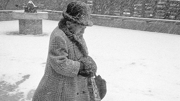 Alte Frau läuft mit Hut und Mantel mit viel Schnee von oben.