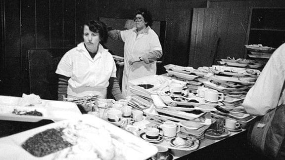Frauen in einer DDR-Großküche stehen vor einem Stapel schmutzigen Geschirrs.