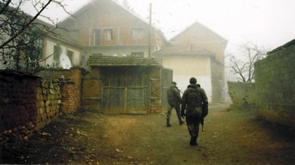 Zwei Soldaten gehen an aufgeschlichtetem Holz auf ein Haus zu