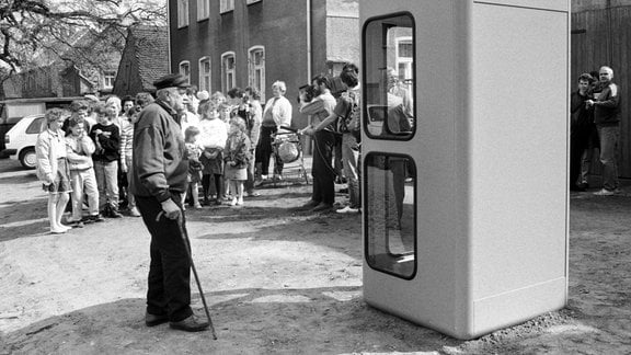 Alter Herr an Krückstock geht zu Telefonzelle, hinter der sich Menschen wie zu einem Gruppenfoto aufgereiht haben.