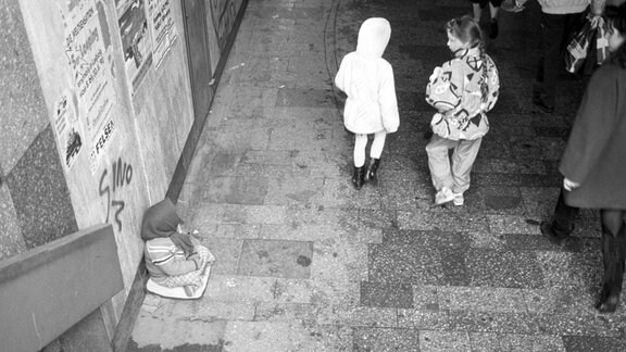 Blick in eine Passage. Links sitzt ein kleines Mädchen am Boden und bittet um Almosen. Rechts gehen zwei Kinder vorbei, eines schaut auf das bettelnde Kind.