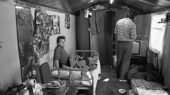 Blick in einen Wagencontainer: Eine Herdplatte, Plakate an den Wänden, ein Bett, zwei Männer, einer der Kamera abgewandt.