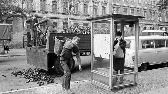 Eine Frau in einer Telefonzelle hält sich ein Ohr zu, weil draußen Kohlen von einem Laster geschaufelt werden.