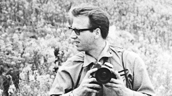 Fotograf Billhardt mit umgehangener Kamera in einem Feld, im Hintergrund Zugwaggons