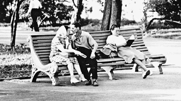 Drei Menschen auf einer Parkbank, links ein Pärchen,d ass zum kusse ansetzt, rechts daneben lesend, eine Frau mit langem Rock