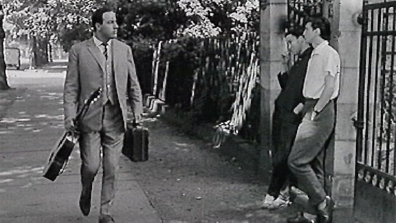 (Filmszene) Manfred Krug spaziert mit Gitarre und Koffer in der Hand an Pärchen in Toreingang vorüber.