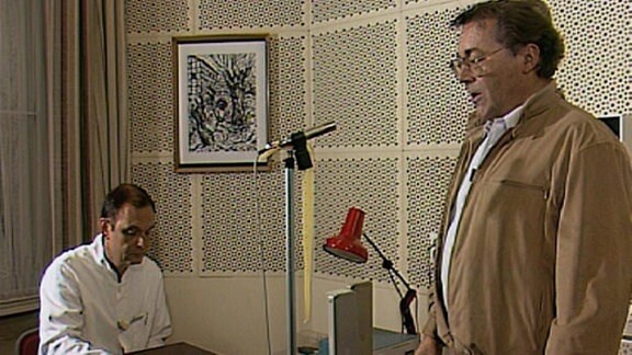 Ein Mann steht vor einem Mikrofon, ein anderer sitzt an einem Aufnahmegerät