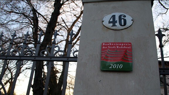 Ein Pfosten, auf dem eine Hausnummer und eine Plakette angebracht sind.