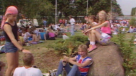 Publikum vor dem Konzert auf einer Wiese: Ein Mädchen in kurzen Hosen sitzt auf einem großen Stein, unter ihr auf dem Rasen sitzt ein Jugendlicher mit Bierflaschen