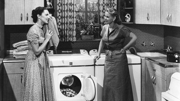 Zwei Hausfrauen vor einer Waschmaschine in einer Küche.