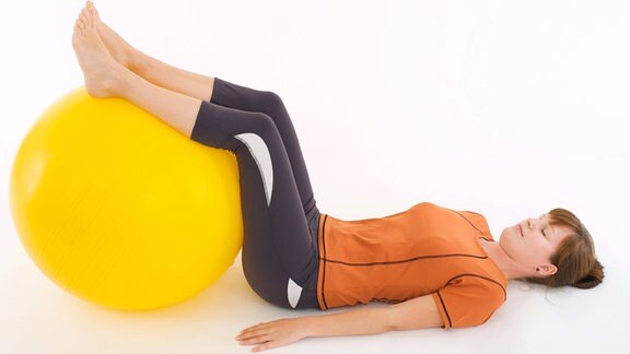 Eine Frau liegt rücklings auf dem Boden und lagert ihre Beine auf einem gelben Gymnastikball