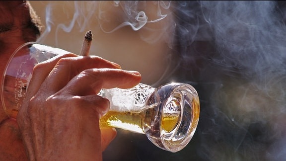 Ein Mann trinkt Bier aus einem Glas und hält dabei eine Zigarette zwischen den Fingern.)