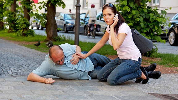 Eine Frau kniet neben einem Mann, der einen Herzinfarkt erlitten hat, und ruft über ihr Mobiltelefon Hilfe
