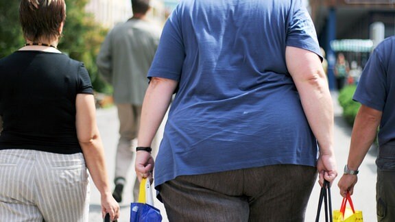 Ansicht von hinten: Zwei übergewichtige Frauen, eine davon stark, laufen in einer Fußgängerzone entlang.