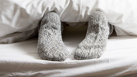Die in dicken Wollsocken steckenden Füße einer im Bett liegenden Frau, schauen unter der Bettdecke hervor.