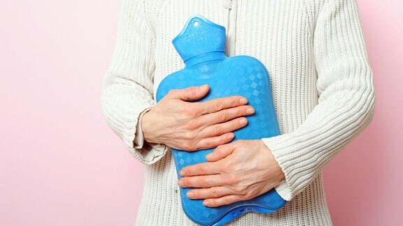 eine ältere Frau in einer weißen Strickjacke hält sich eine blaue Wärmflasche vor den Bauch