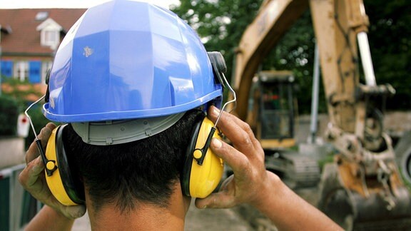 Ein von hinten zu sehender Bauarbeiter, der einen Bauhelm und einen Gehörschutz trägt, steht vor einem Kran.