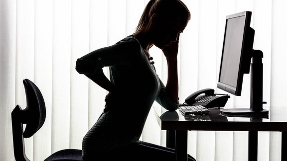 Silhouette einer Frau, die in einem Büro am Schreibtisch sitzt und sich an den Rücken fasst.