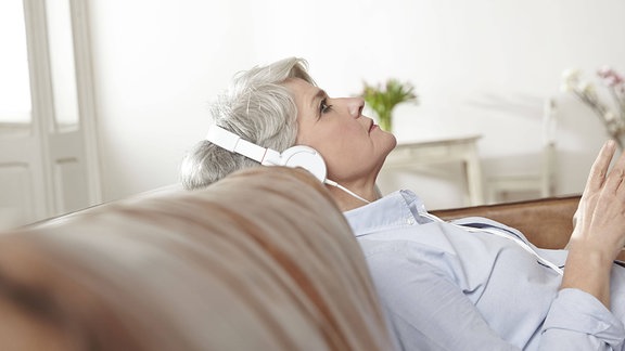 Eine Frau reiferen Alters sitzt in enspannter Position mit Koüfhörern auf einem Sofa und hört konzentriert Musik. 