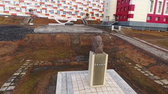 Lenin-Denkmal und Schriftzug "Unser Ziel - der Kommunismus" vor saniertem rot-weiß-gelben Plattenbau in tundraartiger Landschaft
