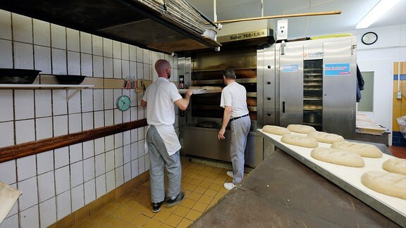 Zwei Männer schieben mit einer Art Brett Brotteiglinge in einen Backofen.