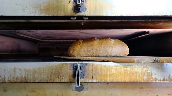 Blick in den schmalen Schub eines Backofens. Vorne liegt ein knusprig braunes Brot auf einem Holzschieber.