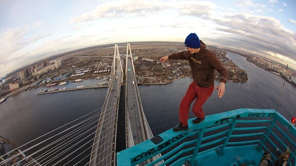 Junger Mann balanciert auf einem blauen Geländer hoch oben über einem Fluss mit einer doppelten Schrägseilbrücke mit jeweils vier Fahrspuren