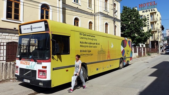 Frau läuft vor einem gelben Bus durch eine sonnige Straße der kosovarischen Hauptstadt Pristina. Auf dem Bus steht: "EU-Quiz and Games". 