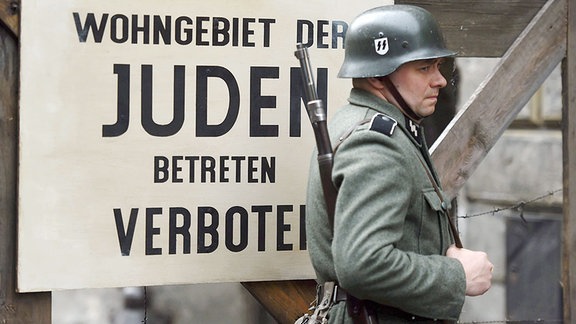 Ein Statist, der ein Mitglied der SS spielt, steht im Filmset vor einem Schild mit der Aufschrift 'Wohngebiet der Juden. Betreten verboten'.